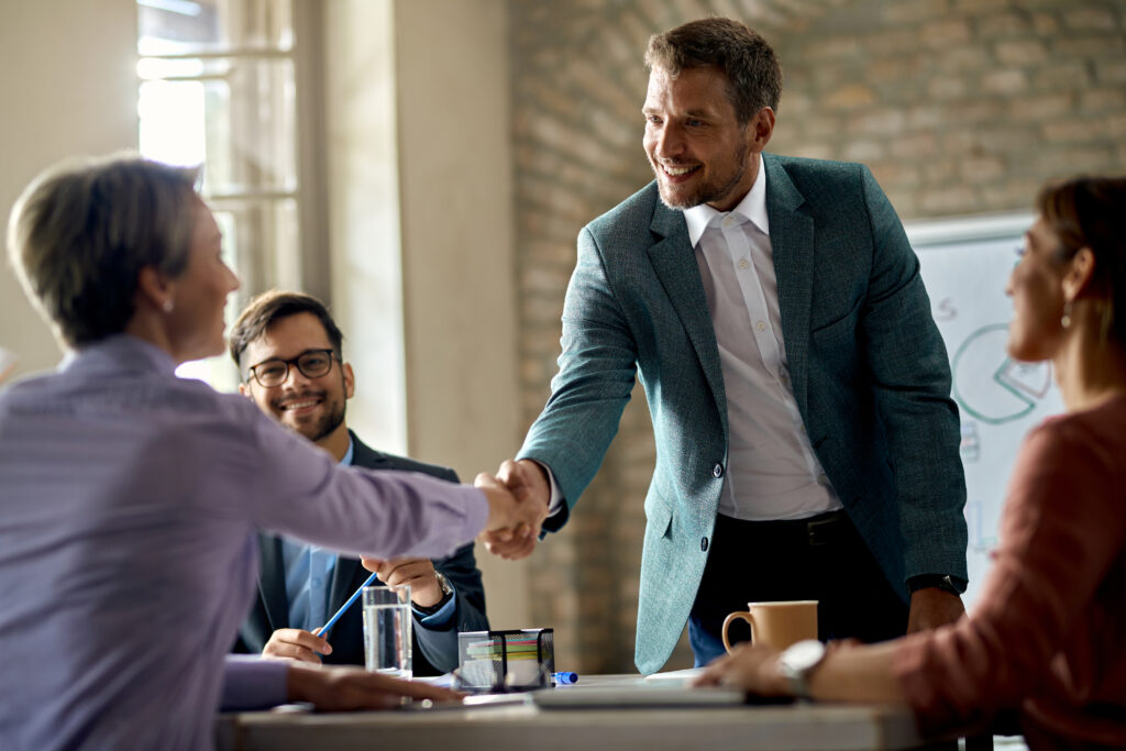 Two professionals shaking hands across a conference table while colleagues look on during a business meeting
